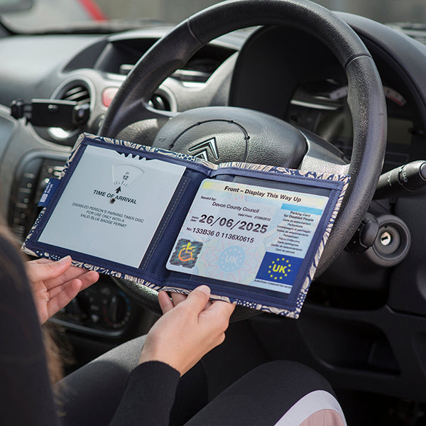 Open Disabled Blue Badge Wallet held in two hands against a steering wheel. The adjustable timer display clock is on the left. On the right is a Devon County Council Disabled Parking Permit with hologram. The acetate window doesn't cover the hologram.