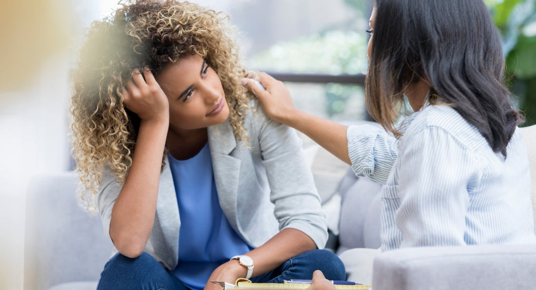 A distressed woman rests her head on her hand while a therapist places a comforting hand on her shoulder。