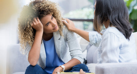 A distressed woman rests her head on her hand while a therapist places a comforting hand on her shoulder。