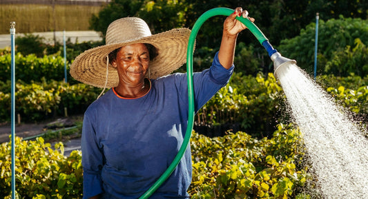 A woman wearing a straw hat and a blue shirt watering plants with a green hose.