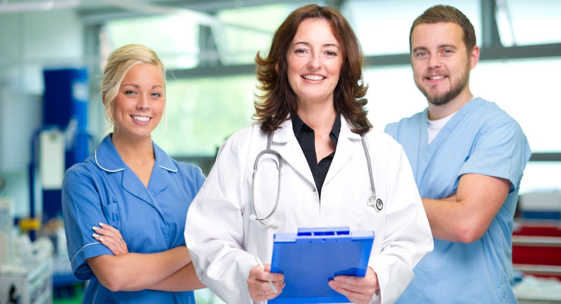 Three smiling healthcare professionals standing together in a clinic; a female doctor in a white lab coat holding a blue clipboard stands in the center, flanked by a female nurse in a blue tunic and a male healthcare worker in light blue scrubs.