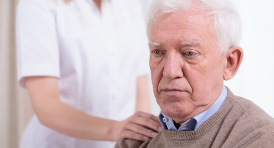 Older man looking solemn while a caregiver in a white uniform rests a hand on his shoulder.