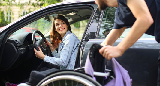 shows a woman in the driver seat of a car smiling back at a person holding a wheelchair outside the open rear door.
