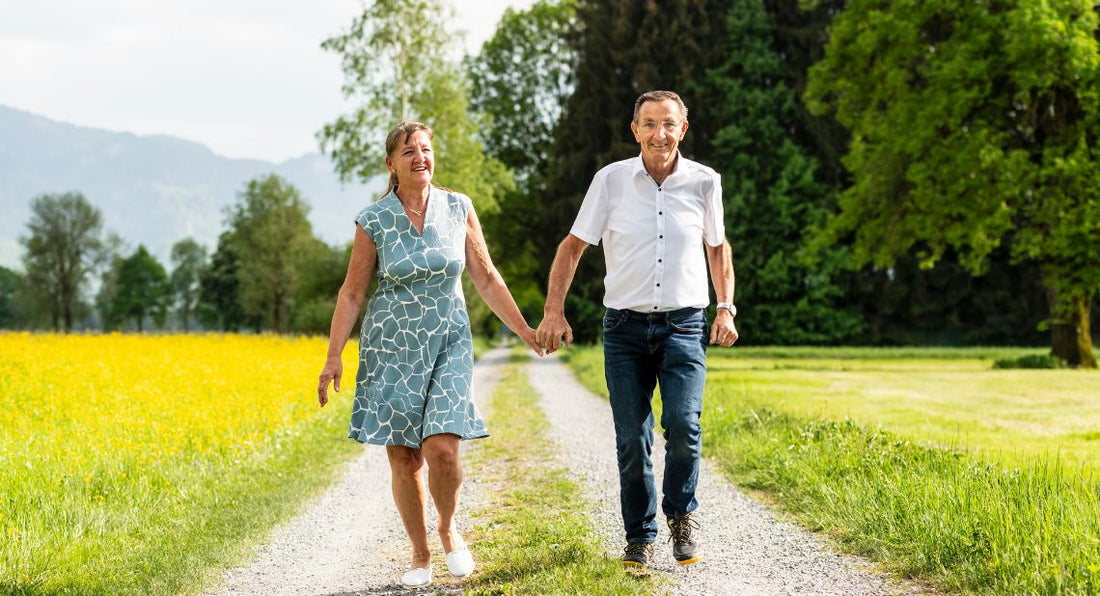 Older couple holding hands while walking down a gravel path next to a field of yellow flowers.