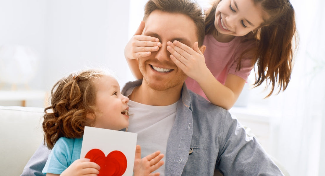 A smiling man sits while an older girl covers his eyes from behind. A younger girl stands in front holding a card with a red heart.