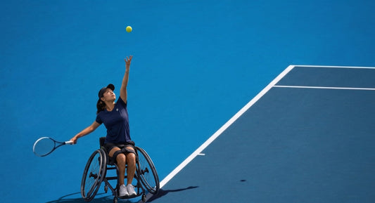 A female wheelchair tennis player serving a ball on a blue court.