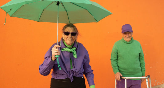 An elderly woman wearing purple holds a green umbrella while standing near an elderly man with a walker against an orange wall.