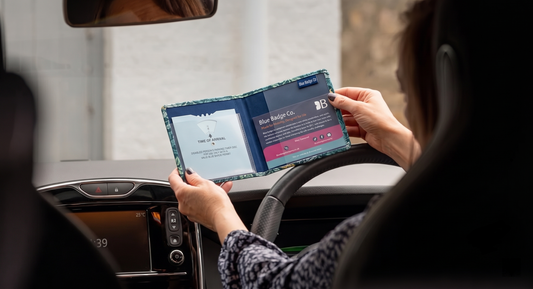 a person in a car holding an open patterned wallet displaying a disabled parking arrival clock and a Blue Badge Co card.