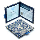 Two Disabled Blue Badge Wallets in William Morris Marigold Indigo fabric are pictured. One stands open showing clock display on left behind plastic window, with room to adjust time dial. On the right is a card insert showing place for permit with a hologram safe design. Second, closed wallet lays in front of the open wallet to display fabric. The William Morris Marigold Indigo fabric is a botanical flower and leaf pattern with cream flowers on a navy blue background.