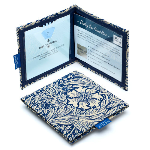 Two Disabled Blue Badge Wallets in William Morris Marigold Indigo fabric are pictured. One stands open showing clock display on left behind plastic window, with room to adjust time dial. On the right is a card insert showing place for permit with a hologram safe design. Second, closed wallet lays in front of the open wallet to display fabric. The William Morris Marigold Indigo fabric is a botanical flower and leaf pattern with cream flowers on a navy blue background.