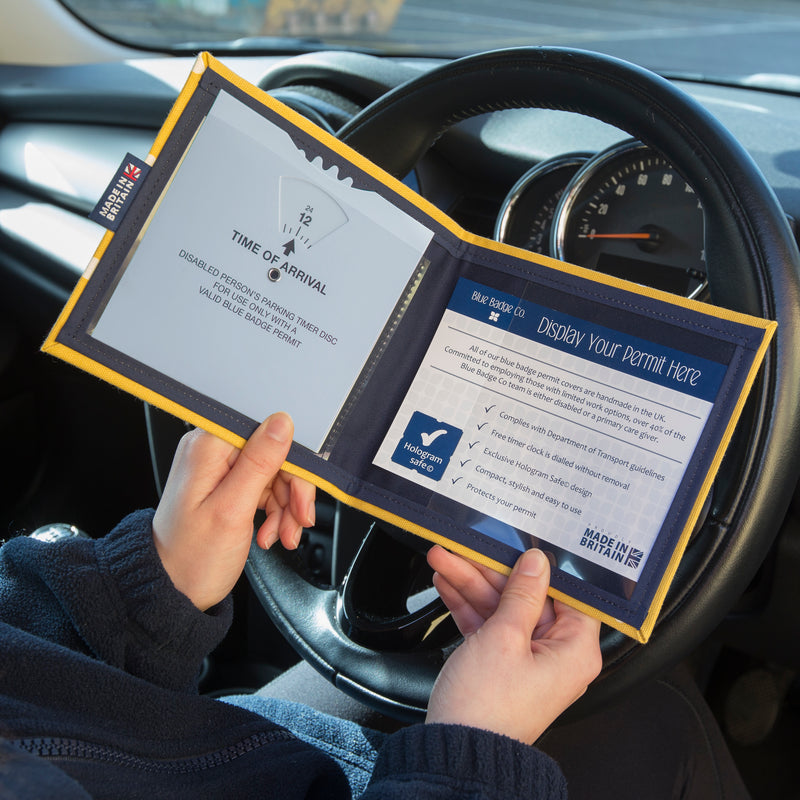 Open Disabled Blue Badge Wallet held in two hands against a steering wheel. The adjustable timer display clock is on the left. On the right is the Blue Badge Company insert with a "hologram safe" box to show how the permit should be displayed. A label half way down the left side of the wallet reads "Made In Britain".