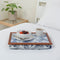 The lap tray in Peacock fabric is pictured on top of a bed dressed with white bed linen. The tray holds a white mug of coffee and a breakfast bowl with fruit in it. A plant stands on a side table next to the bed.