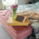 The Canary Yellow Spotty Lap Tray is pictured on a pink velvet ottoman at the foot of a bed. On the tray there is an open book and a vase of pink tulips and hyacinths.