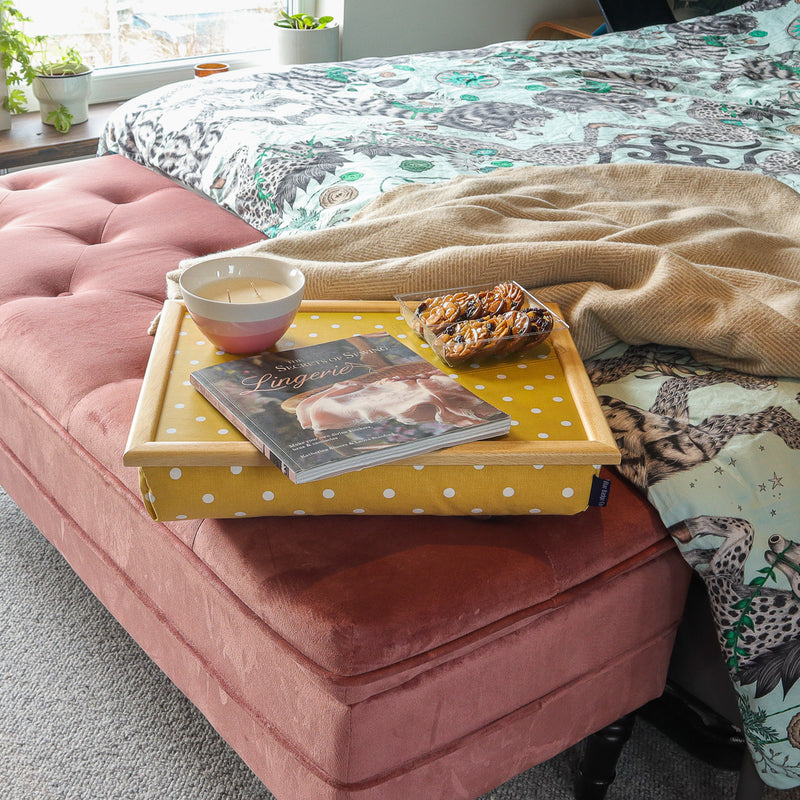 The Canary Yellow Spotty Lap Tray is pictured on a pink velvet ottoman at the foot of a bed. On the tray there are biscuits, a magazine and a bowl candle. 