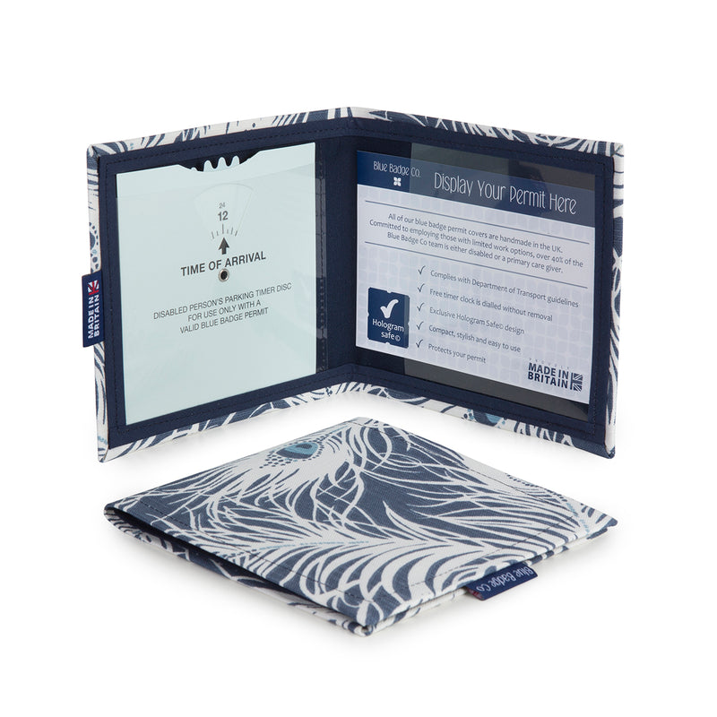 Two Disabled Blue Badge Wallets in Peacock fabric are pictured. One stands open showing clock display on left behind plastic window, with room to adjust time dial. On the right is a card insert showing place for permit and a hologram safe design. Second, closed wallet lays in front of the open wallet to display fabric. The Peacock fabric has a navy blue background with white wisps of the peacock feathers. The centre of the peacock feather is a lighter blue, defining the eye of the feathers classic design.