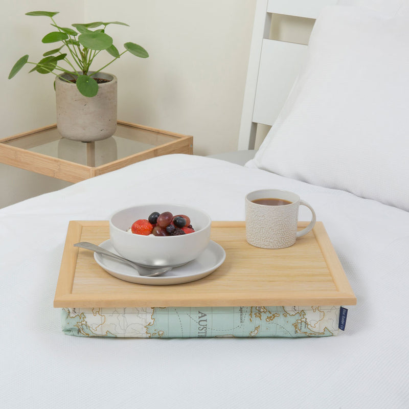 The lap tray in World Map is pictured on top of a bed dressed with white bed linen. The tray holds a white mug of coffee and a breakfast bowl with fruit in it. A plant stands on a side table next to the bed. 