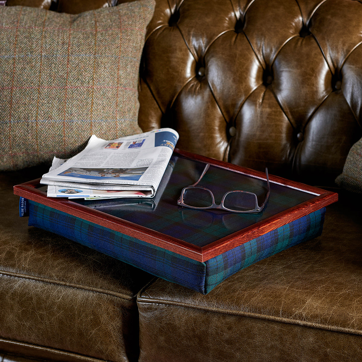 The bean bag lap tray in Blackwatch Tartan is pictured on a brown leather sofa with glasses and a newspaper on top.