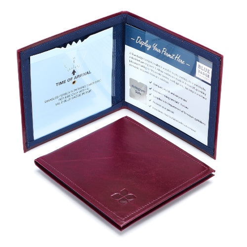Two Disabled Blue Badge Wallets in Burgundy leather are pictured. One stands open showing clock display on left behind plastic window, with room to adjust time dial. On the right is a card insert showing place for permit and a hologram safe design. Second, closed wallet lays in front of the open wallet. The leather is a rich burgundy colour embossed in the bottom right corner with a discreet Blue Badge Co logo.
