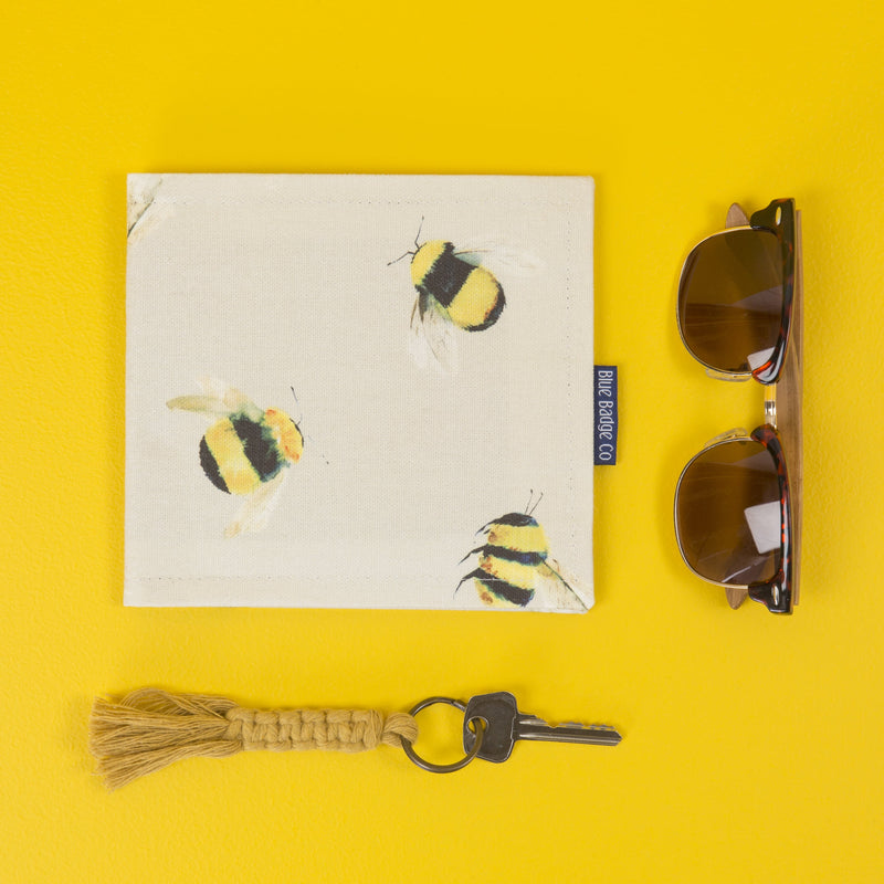 The Blue Badge wallet in Busy Bees fabric lays flat on a yellow surface. To right, a pair of sunglasses, below a small key on a knotted yellow cord key fob.