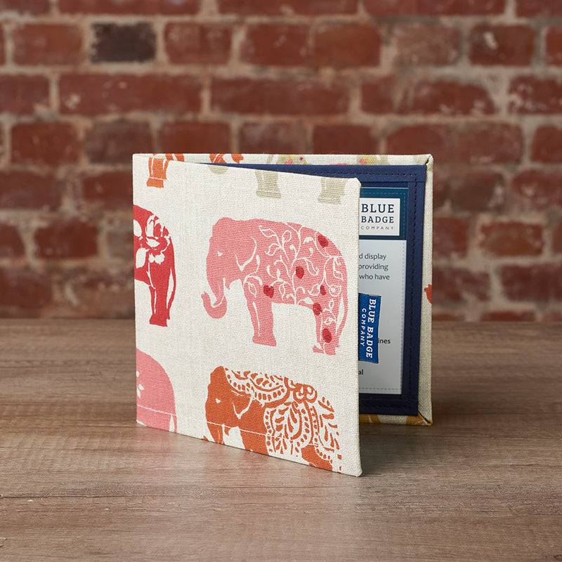 Disabled Blue Badge Wallet in Nelly Elephant fabric is standing slightly open on a wooden surface in front of a brick wall.