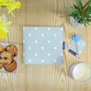 The Blue Badge Wallet in Aqua Marine Spotty is viewed from above as it lays closed on a wooden table along side a RADAR key with a blue rubbery top and matching Aqua Marine Spotty square fabric key fob. A plant is in the top right, daffodils top left. A candle bottom right and some Florentine biscuits bottom left.