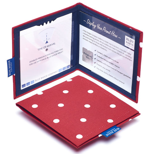 Two Disabled Blue Badge Wallets in Red Spotty fabric are pictured. One stands open showing clock display on left behind plastic window, with room to adjust time dial. On the right is a card insert showing place for permit with a hologram safe design. Second, closed wallet lays in front of the open wallet to display fabric. The Red Spotty fabric is a deep red with white spots dotted in a regular symmetrical pattern, an inch apart.