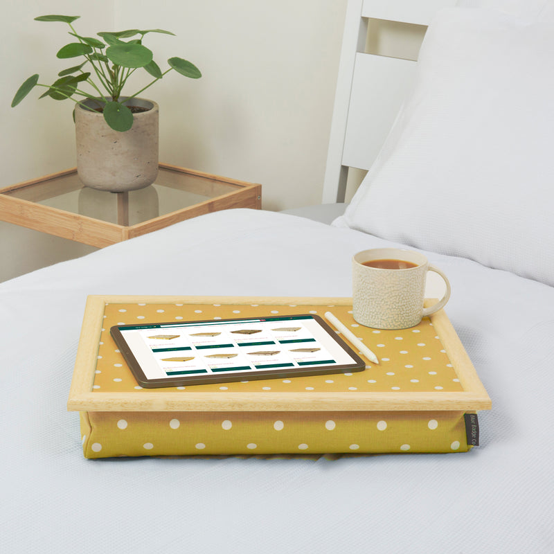 The bean bag lap tray in Canary Yellow Spotty is pictured on top of a bed dressed with white bed linen. The tray holds a white mug of coffee, an electronic tablet and a pen. A plant stands on a side table next to the bed.