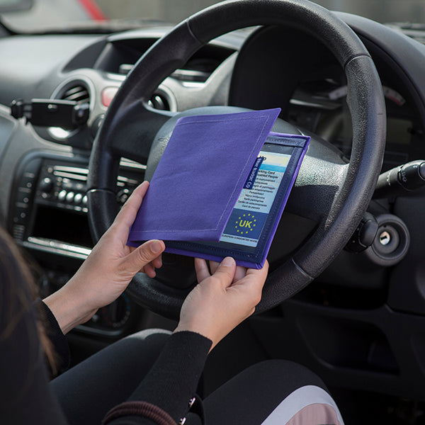 The Blue Badge Wallet is held slightly open, in two hands, against a steering wheel.