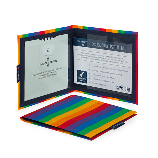 Two Disabled Blue Badge Wallets in Rainbow fabric are pictured. One stands open showing clock display on left behind plastic window, with room to adjust time dial. On the right is a card insert showing place for permit and a hologram safe design. Second, closed wallet lays in front of the open wallet to display fabric.  The Rainbow fabric has bright bold stripes in the traditional rainbow colours of red, orange, yellow, green, blue and purple.